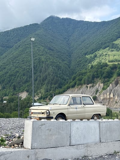 An abandoned, light-colored sedan sits behind a concrete barrier. The car is a Soviet-era ZAZ-968M, identifiable by its distinctive rear engine air vents and headlights. It is situated in a rural or mountainous area, with a lush green mountain range in the background. The sky is overcast, and a street light is visible on a pole to the left of the car. The foreground is filled with concrete blocks and gravel. The scene appears to be located in or near Kvesheti, Georgia, a region known for its mountainous terrain and Soviet-era infrastructure.