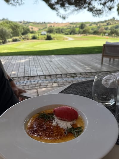 A close-up shot focuses on a white dessert bowl containing a crème brûlée topped with a scoop of pink sorbet, a sprig of rosemary, and a dusting of powdered sugar. The dessert rests on a dark grey placemat on a wooden deck. In the background, a lush green golf course stretches out towards rolling hills covered in vineyards and trees under a clear sky. A small white building is visible in the distance on the hillside. Part of a person's arm and hand are visible on the left side of the frame, resting on the edge of the table. Two empty drinking glasses and a white napkin are also present on the placemat. The scene appears to be at a restaurant or cafe with an outdoor seating area overlooking a golf course, likely located in the vicinity of Torres Vedras, Portugal, known for its scenic golf resorts and wine regions.