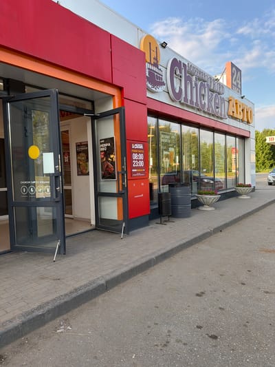 The image shows the exterior of a "Chicken ABTO" fast-food restaurant located in Krilovo, Russia. The building features a bright red facade with large glass windows and automatic doors that are partially open. A prominent sign displays "SOUTHERN FRIED Chicken ABTO" in large, multi-colored lettering. The restaurant's operating hours are displayed on a red board: "РЕЖИМ РАБОТЫ 08:00 - 23:00".

On the sidewalk in front of the restaurant, there are two decorative planters filled with pink flowers. A dark gray trash can is situated near the entrance. Through the glass windows, reflections of trees and passing cars are visible. The sidewalk is paved with brick pavers, and the street in front of it is asphalt. The sky is partly cloudy, suggesting it is daytime. There are no people visible in the immediate vicinity of the restaurant entrance or the surrounding area. The overall scene depicts a typical commercial establishment on a street in a Russian town.