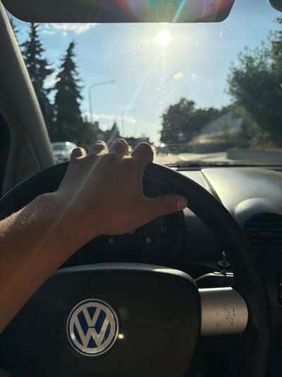 This media file shows a person's hand on the steering wheel of a Volkswagen car, viewed from inside the vehicle. The sun is shining brightly through the windshield, creating lens flares and illuminating the interior. The scene appears to be a road, with trees and the sky visible through the windshield. The driver's hand is positioned at the top of the steering wheel, suggesting they are actively driving. A Volkswagen logo is clearly visible on the airbag cover. The bright sunlight suggests it is daytime, likely a sunny day. The context provided places the location as Brno, Czechia. No specific emotions are conveyed by the image, other than perhaps a sense of being on a journey. There is some text visible on a sticker on the left side of the windshield, but it is largely illegible due to glare and resolution.