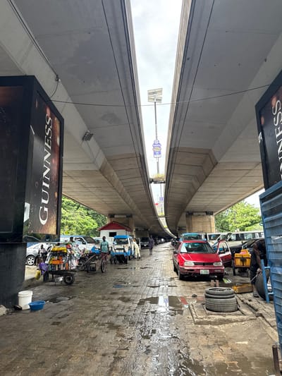 This is a ground-level, eye-level, medium shot of a busy street under an elevated highway in Ikoyi, Nigeria. The concrete structure of the highway dominates the upper portion of the frame, with its underside casting shadows on the scene below. A large, vertical Guinness advertisement is prominently displayed on the left, with text running vertically.
The street itself is paved with interlocking bricks and is wet and reflective, indicating recent rain or a street cleaning operation. Several vehicles are present, including a red car parked in the foreground with its door open, a blue and white minibus, and other cars and vans further down the street. There are people on foot, some appearing to be vendors with carts piled high with goods, and others walking or tending to vehicles.
On the right side of the frame, a man is shown working on tires, with a stack of tires next to him and a yellow container. The environment appears to be a commercial area with a mix of vehicular traffic and pedestrian activity. The overall impression is one of a lively, urban street scene.