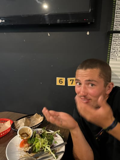 The image depicts an indoor scene in Wombarra, Australia, likely a casual dining establishment or pub. A young adult male with short brown hair and a faint mustache is seated at a wooden table, looking directly at the camera with a slightly open-mouthed expression and his left hand raised near his chin, palm forward. He wears a dark short-sleeved shirt and a blue and multi-colored bracelet on his right wrist. A thin ring is visible on his left ring finger. On the table, a white plate holds the remnants of a salad, consisting of mixed greens, a tomato slice, and a small plastic cup with brown dressing residue. A fork and knife are on the plate. Adjacent to the plate are a black plastic basket and a red plastic basket, both lined with brown paper. The background features a dark wall. Mounted high on the upper left portion of the wall is a black LG television screen. To the mid-right, two yellow square plaques with black numbers "6" and "7" are affixed to the wall. Further to the right, a whiteboard displays a vertical list of handwritten names and numbers, including entries such as "Ellie," "Sean," "Dirie," and "Manny."