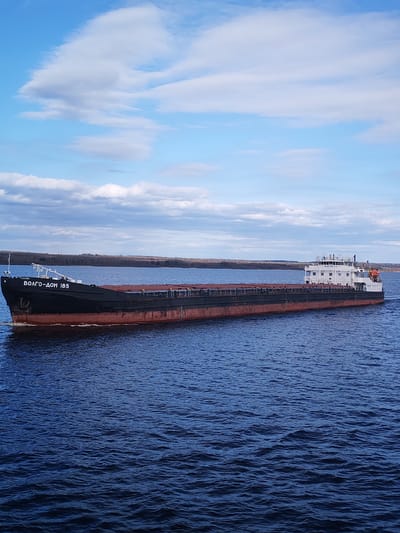The image shows a large cargo ship, identified as "ВОЛГО-ДОН 185" (Volgo-Don 185), navigating a wide body of water under a partially cloudy blue sky. The vessel is a dark-hulled bulk carrier with a reddish-brown waterline, appearing to be laden with cargo, possibly gravel or another bulk material, visible in its open holds. Its superstructure, located at the stern, is white and features multiple levels, windows, and navigation equipment, including an orange life raft mounted on the side. The ship is moving from left to right, creating a distinct wake behind it and at its bow.

The water is a deep blue, showing ripples and small waves. In the distance, a low, forested shoreline stretches across the horizon, suggesting a large river or canal. The sky above is clear blue in many parts, with scattered white and light gray cumulus clouds. The overall lighting indicates it is daytime, likely late morning or early afternoon, with clear visibility.

The most prominent text is "ВОЛГО-ДОН 185" painted in large white Cyrillic letters on the ship's black bow. A smaller number, "130364," is also visible near the bow above the main text. The name "Volgo-Don" is indicative of its operation in the Volga-Don region of Russia, consistent with the provided location context of Novaya Svobodka, Russia. The scene portrays routine commercial navigation on an inland waterway. No individuals are clearly visible on the ship or elsewhere in the frame.