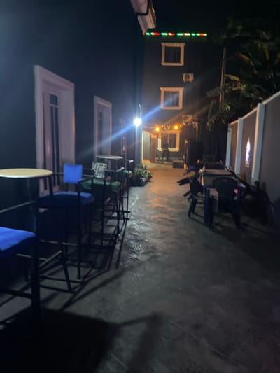 The scene depicts an outdoor commercial establishment at night in Igbessa, Nigeria. The area is illuminated by multiple artificial light sources, casting strong glare and elongated shadows on the wet or reflective paved ground.
On the left, a dark-colored building facade features two tall, white-framed vertical openings. In the foreground-left, a row of five dark-framed bar stools with blue upholstered seats is positioned next to a round, light-colored table. Further back on the left, additional tables and chairs, including one with a striped cushion, are present.
In the background, a multi-story, dark-colored building is visible, featuring two rectangular white-framed windows and two exterior air conditioning units on its upper facade. The building's top edge is adorned with a line of red and green decorative lights. A brightly lit entrance, framed by string lights with warm yellow bulbs, is visible at the ground level of this building. A solitary, indistinct human figure is partially visible within this entrance.
On the right, a dark gray wall or structure exhibits a repetitive pattern, including a design resembling a stained-glass window. Several dark-colored plastic chairs and tables are irregularly arranged on the right side of the paved area, some appearing stacked. Sparse green foliage is visible adjacent to the background building.