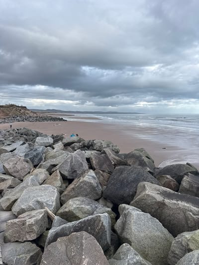 The image displays a coastal scene in Montrose, United Kingdom, under a heavily overcast sky composed of dark grey clouds. The foreground is dominated by a large, irregular barrier of angular grey and dark grey boulders serving as a sea defense. A single piece of light-colored driftwood is visible among these rocks.

To the left, beyond the immediate rock barrier, a section of sandy beach transitions into vegetated sand dunes topped by a faint linear structure, possibly a fence or pathway. An individual wearing a light-colored top and dark trousers is visible walking from right to left on the wet sand at the base of these dunes, positioned further down the coastline.

In the midground, closer to the rock barrier, another individual wearing a bright blue top is observed on the wet sand, seemingly seated or kneeling, with a small, dark-colored animal positioned next to them.

The majority of the midground consists of a broad, flat expanse of wet, light brown sand, indicating a receding tide or low water level. This sand reflects the dull light from the overcast sky.

The right side and background feature the North Sea, characterized by gentle waves breaking on the shore, its surface reflecting the grey sky. The distant horizon reveals a subtly curved coastline with faint landmasses.