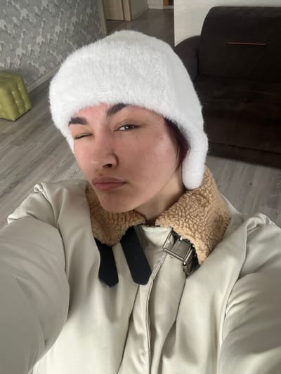 This image depicts a single individual within an indoor residential setting in Starobaltachevo, Russia. The individual, appearing to be an adult female, is centered and occupies the majority of the frame, captured in a selfie-style, upward-angled perspective. She has dark hair, dark eyebrows, and her left eye is closed in a distinct wink, while her lips are pursed forward. She is wearing a white, fluffy winter hat, covering her head and ears. Her attire also includes a light-colored, possibly off-white or beige, puffer jacket with a prominent light brown, sherpa-style collar. A metallic silver-colored buckle and two black strap elements are visible on the right side of the jacket's chest area. The background elements include a light wood-toned floor. To the left, a wall is covered with a light gray, abstract, cloud-like patterned wallpaper, with a light green upholstered cube or ottoman resting on the floor against it. To the right, a section of a dark brown upholstered sofa is visible. A doorway with a light-colored frame is partially visible in the upper left background. No other individuals or interactions are depicted.