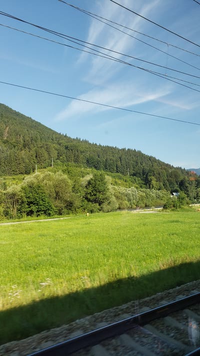 The image captures a scenic view from a moving train, likely in or around Stankovany, Slovakia. The foreground features a blurred view of train tracks and ballast, indicating the perspective of a passenger. A shadow is cast across the vibrant green field in the middle ground, suggesting the sun is positioned behind the viewer. The field is dotted with wildflowers. Beyond the field, a dense, verdant forest covers the slope of a large hill. A winding road is visible at the edge of the field before it meets the trees. In the distance, a body of water, possibly a river, can be seen. The sky above is a clear blue, with wispy white clouds streaking across it. Several overhead power lines traverse the top of the frame. A few utility poles are scattered along the hillside within the treeline. A small structure, likely a building, is partially visible nestled amongst the trees on the right side of the hill. The overall scene is one of natural beauty and tranquility, characteristic of rural landscapes.
