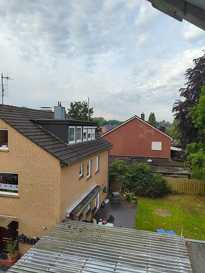 The image depicts a residential area in Papenburg, Germany, on an overcast day. In the foreground, a corrugated metal roof is visible, showing signs of weathering. Beyond this, a yellow brick house with a dark tiled roof extends to the left. This house features several windows and a dormer with multiple windows. To the right of this house, a larger red brick building is partially visible, with a small white-shuttered window on its side. A backyard area with a patio, lawn, fence, and various objects including a grill and lounge chairs is situated behind these houses. Lush green trees and shrubs surround the property, with a particularly dark-leaved tree prominent on the right side of the image. The sky is filled with light grey clouds. No people are present in the scene.
