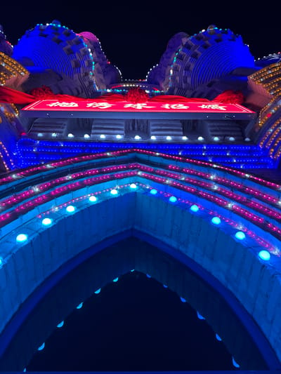 A nighttime, low-angle view captures a brightly lit traditional Chinese architectural structure in Baqiao, China. The structure features an archway with rows of spherical blue lights outlining its curves. Parallel to the archway are vertical lines of pink and blue lights. To the right is a red sign with white Chinese characters, adorned with fabric decorations. The building is elaborately tiered and illuminated with blue and gold lights. The background is a dark night sky.