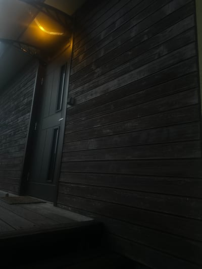 The image shows a dark wooden exterior wall with horizontal planks, part of a building in the city of Lapės, Lithuania. In the foreground, a dark gray door is visible, flanked by narrow vertical windows. Above the door, an awning with a bright yellow light illuminates the entrance. To the left of the door, a small, dark doormat rests on a wooden porch. The overall scene is dimly lit, suggesting it is either dusk or nighttime.