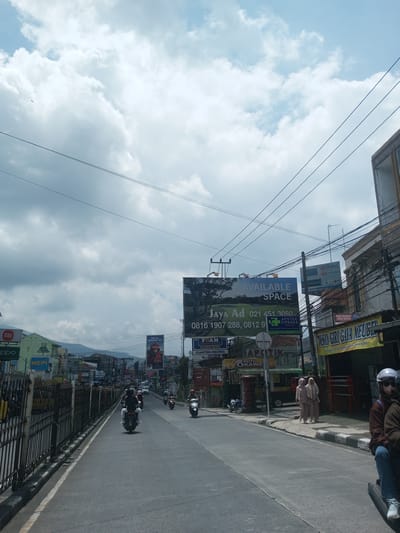 The image shows a street scene in an urban area, likely Southeast Asia based on architectural style and the presence of individuals in hijabs. The street is paved and has a divider separating traffic. Motorcycles are present on the road and parked along the side. A large billboard advertising "AVAILABLE SPACE" is prominent. Buildings line the street, housing various businesses identified by signage in what appears to be an Indonesian/Malay language, including "APOTIK" (pharmacy) and "TOKO GIRI GAYA MEUBEL" (furniture store). Two women wearing hijabs are walking on the sidewalk. The sky is overcast with clouds. It cannot be precisely identified in Xayabury District, Laos, because signs are in Indonesian/Malay.