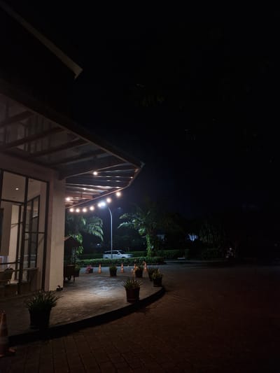 The image shows a nighttime scene in Banyuwangi, Indonesia. A building with a modern, partially exposed wooden frame roof is visible, adorned with a string of lights. The building has large windows, some of which are illuminated. A pathway lined with potted plants and trees leads away from the building. A street lamp illuminates the area. The pathway appears to be made of paving stones. The overall scene is dark, with artificial lighting providing the primary illumination.