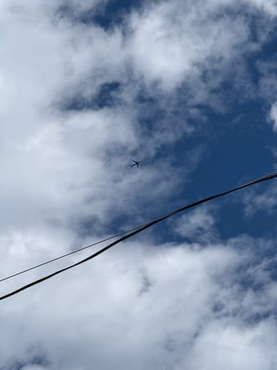 An airplane is visible in the upper portion of the image, flying against a backdrop of a deep blue sky filled with white, fluffy clouds. Two black wires traverse the image diagonally from the lower left to the upper right, obscuring parts of the sky and clouds. The overall scene appears to be outdoors, under daylight conditions. The location is identified as Дзержинец-83 Снт, Russia.
