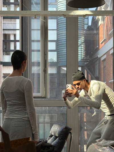 The image captures an indoor setting, likely a residential space, within the city of Saint-Denis, France. Two individuals are present in a brightly lit room, illuminated by strong natural sunlight entering through a large multi-pane window that frames an urban exterior.

Subject 1, female, is positioned on the left, facing away from the viewer, looking towards the window. She has short, dark, slicked-back hair and wears a sheer, light-colored long-sleeved top over a darker undergarment, paired with light grey trousers.

Subject 2, male, occupies the right side, observed in a crouched or bent-over stance. He wears a dark head covering (durag or similar), a light-colored, long-sleeved top with a V-neck, and light grey sweatpants with visible "adidas" branding on the left leg. He holds a white smartphone horizontally with both hands, actively directing its camera towards Subject 1 or the exterior through the window.

Essential objects within the room include a dark, tufted recliner-style chair with metallic legs situated centrally, a partially visible brown container (possibly a bag or box) in the lower-left foreground, and light fabric with plastic packaging in the lower-right foreground. A dark, bell-shaped light fixture hangs from the ceiling, partially visible at the top.

The exterior view through the window reveals a diverse urban landscape: modern structures with reflective windows on the left, contrasting with older red-brick buildings featuring traditional windows and a distinctive cylindrical, ribbed structure on the right.