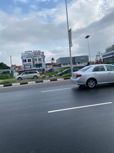 This image depicts a street scene in Ojomu, Nigeria, on an overcast day. The foreground shows a dark asphalt road with two solid white lane markings. The road surface appears wet or reflective. A raised median strip, edged with a black and white striped curb, separates the traffic lanes.
On the median, a light green minivan, identifiable as a Toyota Sienna, is parked facing left. Behind it, a dark-colored sedan, possibly a Hyundai Elantra, is also parked, partially obscured. A single individual wearing dark clothing is visible near the rear of the dark sedan, moving away from the viewer.
Dominating the background on the left is a two-story building named "ALPHA PLAZA," prominently displayed in large white letters. Below the plaza name, a sign reads "NOW LETTING" followed by two phone numbers: "0806 412 4226" and "0806 284 5290." The first floor features a business called "KIDS DESIGN COMPANY." The ground floor displays "LaLEKSIDE" and "TRINI Y DOORS" as storefronts. Adjacent to Alpha Plaza, to the left, are older buildings with reddish roofs amidst green foliage.
Further down the road, to the right of Alpha Plaza, a modern building with dark grey or blue cladding is visible. A tall utility pole with multiple cross-arms and power lines stands near Alpha Plaza. In the mid-ground, a silver Toyota Corolla sedan is observed on the road, partially facing left. Its rear bumper and side profile are visible, including its alloy wheels.
To the right of the frame, a tall, slender metal pole features a dark red or maroon banner, mostly unreadable. Another similar pole, topped with a solar panel, indicates a street light. The sky is heavily clouded with a mix of grey and white, suggesting an overcast day with no direct sunlight.