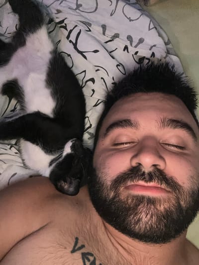 This is a close-up, overhead shot of a person sleeping on a bed, with a black and white cat nestled beside them. The person, who appears to be male, has their eyes closed and a slight smile on their face. They have dark hair, prominent eyebrows, and a full beard. A tattoo, partially visible on the person's chest, contains the letters "V." The cat is curled up, appearing to be asleep as well, with its head resting on the person's arm or chest. The bedding is white with a subtle black abstract pattern. The lighting suggests it could be daytime or indoor lighting. The overall mood is peaceful and intimate, conveying a sense of comfort and companionship.