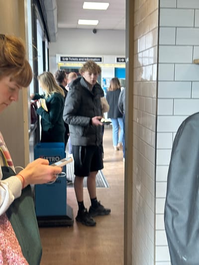 The image appears to be taken indoors, likely in a subway station in Tonbridge and Malling, United Kingdom. Several people are visible, primarily standing in line. One person is holding a phone, possibly taking the picture. Another individual is wearing a dark grey jacket, black shorts, and black shoes. A blue box with "METRO" printed on it is also in view. The background shows a white tiled wall and a sign that may say "Tickets and Information." The overall environment seems like a typical waiting area at a subway station.