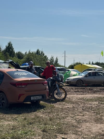 The image depicts an outdoor scene at what appears to be a campground or event in Chyormoz, Russia. In the foreground, an orange Kia Rio sedan is parked on a muddy surface. A sticker on its rear window reads "НАНИМАШ ABTOKУРЬЕРОВ +7707". A motorcycle is parked to the right of the Kia.
In the middle ground, two men are standing and interacting. One man, wearing a red shirt and sunglasses, is gesturing towards something out of frame. The other man, wearing a dark shirt and a bandana, is holding a phone to his ear and wearing sunglasses. Behind them, several cars and tents are visible, including a large yellow tent and a green tent. The environment is characterized by dirt, mud, and sparse vegetation, with trees in the background under a clear blue sky with some wispy clouds. A yellow flag is visible on a pole in the distance. Electrical poles are also present in the background.
