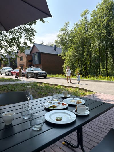 A medium shot captures an outdoor seating area at a cafe in Morozovo, Russia, on a sunny day. In the foreground, a dark-colored table is set with plates, cups, and glasses, some of which appear to be remnants of a meal. Plates contain leftover food, including what looks like cheese, fruit, and pastries. A white cup with a tea bag rests on a saucer. In the middle ground, a paved walkway runs parallel to a grassy area. Several modern brick houses with dark roofs are visible behind the walkway. Two women and a small black dog are walking on the walkway, approaching parked cars. Further down the walkway, a woman in a white dress and a young child in a blue outfit are walking away from the camera. Dense green trees line the right side of the walkway, creating a natural backdrop. The scene is illuminated by bright sunlight.