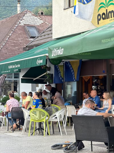 The image depicts an outdoor seating area of a cafe, identified as "Caffe bar La vie" with a Tuborg advertisement, in Travnik, Bosnia and Herzegovina. Several patrons are seated at tables. In the background, a building with a tiled roof and a skylight is visible, along with a green awning extending over the cafe's seating area. Two flags, one appearing to be the flag of Bosnia and Herzegovina, are displayed on the building. Various chairs of different colors and materials are arranged around the tables. Some people are engaged in conversation, while others are looking towards the camera or elsewhere. The ground is paved with patterned tiles.