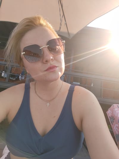 A woman with blonde hair and sunglasses is taking a selfie outdoors. She is wearing a navy blue swimsuit top and a necklace with a cross pendant. The sun is shining brightly, casting a lens flare across the image. In the background, there is a building with a balcony and some laundry hanging on a line. The scene appears to be in Pirogovo, Russia, possibly at a resort or residential area.