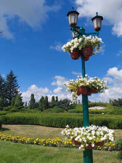 A lamppost adorned with planters of white flowers stands in a park on a sunny day with a blue sky and scattered clouds. The lamppost has three lamps and several tiers of flower pots. In the background, there are manicured green hedges, trees, and a bed of yellow flowers. The location is confirmed to be Kovalivka, Ukraine.