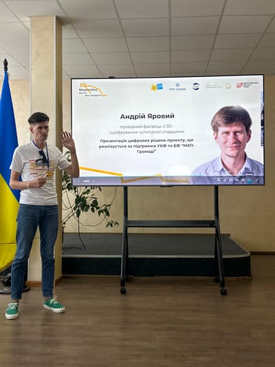 This media file captures a presentation taking place indoors, likely in a community or cultural center, given the presence of a stage-like structure and presentation screen. The main subjects are a man, identified by text on the screen as Andriy Yarovyi, who appears to be the presenter, and a large projection screen displaying information about his project.
Andriy Yarovyi is a young man, dressed casually in a white t-shirt, blue jeans, and distinctive green checkered sneakers. He is holding a golden object, possibly an award or a prop related to his presentation, and gesturing with his other hand. His expression seems engaged and perhaps a little animated, suggesting he is actively addressing the audience.
The projection screen is the focal point of the presentation. It displays a portrait of a man, possibly Andriy Yarovyi himself or a key figure associated with the project. Text on the screen, in Ukrainian, identifies Andriy Yarovyi as a "leading specialist in 3D digitization of cultural heritage" and states that the presentation is about a "digital solutions project, which is being implemented with the support of the Ukrainian Cultural Foundation and the 'Myronivka Community' Charitable Foundation." Several logos are visible on the screen, including those of Myronivka City Council and other supporting organizations. The slide number "16/23" indicates it's part of a larger presentation.
The setting appears to be a room with beige walls and a tiled ceiling. A blue and yellow flag, likely the Ukrainian flag, is partially visible in the foreground. The lighting suggests it's daytime, but the indoor environment makes it difficult to determine the exact time. The overall atmosphere seems to be one of professional engagement and community involvement, centered around a cultural heritage digitization project.