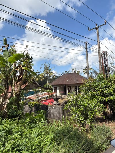 The image shows a rural scene in Susut, Indonesia, with a small house, lush vegetation, and utility poles with overhead wires. The house has a traditional tiled roof and is situated behind a concrete wall. Banana trees and other greenery surround the property. In the background, palm trees are visible, suggesting a tropical climate. The sky is blue with scattered white clouds. The overall impression is one of a peaceful, rural setting.