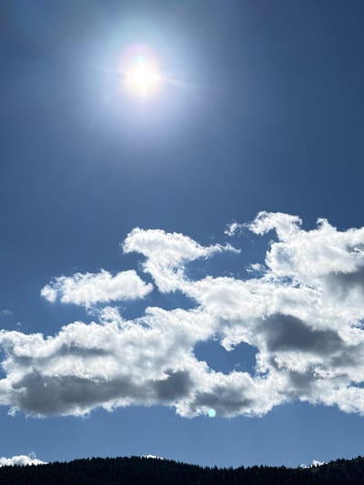 The image depicts a sunny sky with scattered white clouds above a dark, densely forested hillside. The sun, a bright white orb with a distinct lens flare, is positioned in the upper left quadrant of the sky. The clouds are predominantly white and fluffy, with some exhibiting darker, shadowed undersides, suggesting depth and dimension. The forested hillside, appearing dark green to black, forms a solid band across the bottom of the image, its texture rendered as a dense mass of tree tops. The sky itself is a clear, vibrant blue. The scene appears to be an outdoor natural environment. There are no people or animals visible in the image.