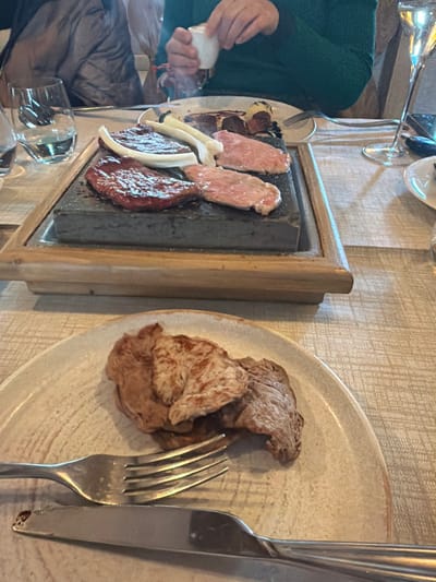 An indoor dining scene in Livigno, Italy, is centered on a hot stone grilling experience. A dark gray rectangular cooking stone, set within a light wooden tray, occupies the mid-ground of a light-colored, patterned tablecloth. On the stone, four pieces of meat are cooking: one deeply seared, dark red cut, and three lighter, pinkish raw slices. Two long, white vegetable segments are also on the stone, with faint steam rising. In the upper right background, a person wearing a green long-sleeved shirt holds a small white pouring vessel above the cooking stone. To the left, a clear drinking glass filled with water is visible, and to the right, a tall, slender champagne flute holds a clear liquid. In the foreground, a light-toned ceramic plate presents several pieces of cooked, browned meat, accompanied by a silver fork and knife. A second person is partially visible in the upper left background.