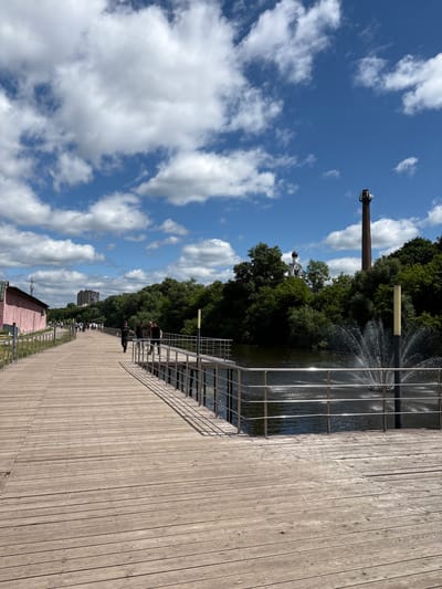 A wooden boardwalk stretches across a body of water, lined with a metal railing. Several people are walking on the boardwalk. On the left, a long pink building is visible. In the background, dense green trees line the water's edge. A tall, brick smokestack stands in the distance, with a church spire to its left. A fountain sprays water in the foreground. The sky is blue with scattered white clouds. This scene is the embankment of the city of Noginsk, Russia.