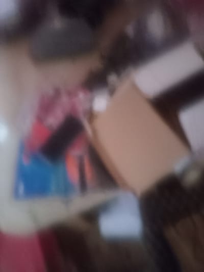 The image, taken in Girei, Nigeria, is a close-up, out-of-focus shot of a table with various items on it. In the foreground, a light brown cardboard box is visible, appearing to be partially open. To the left of the box, a stack of items is discernible, including what looks like a blue rectangular object and some red and black colored shapes. The background is dark and indistinct, suggesting an indoor setting. The lighting is dim, and the overall quality of the image is poor due to the blurriness.