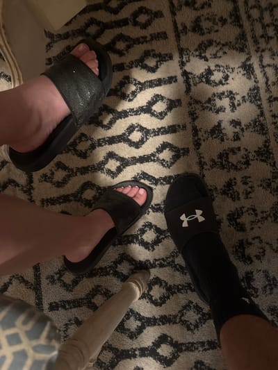 The image shows a close-up of two people's feet resting on a patterned rug. One person is wearing black slide sandals with glitter detailing on the strap, and the other is wearing black slide sandals with the Under Armour logo and black socks. The rug has a cream background with a black abstract geometric pattern. The lighting is dim, suggesting an indoor setting. The image is undated and unlocated, and no specific city or country is mentioned.