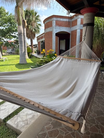 The image depicts a hammock suspended in a garden setting. The hammock is made of light-colored fabric, possibly cotton or a similar material, stretched between two wooden spreader bars. The strings connecting the hammock to the anchor points are visible at one end. The hammock is situated on a tiled patio, with grass and other greenery visible beyond. In the background, there is a brick building with arched windows and a cylindrical architectural element. Palm trees and other trees are scattered throughout the garden. The image suggests a tranquil and relaxing outdoor space in the city of San Francisco Lachigoló, Mexico.