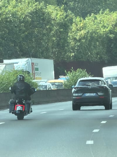 In Savigny-sur-Orge, France, on a highway, a motorcyclist wearing a black leather jacket and helmet rides a black motorcycle with saddlebags. The motorcycle has dual exhaust pipes and a license plate visible. To the right of the motorcycle, a dark-colored Mazda hatchback with a visible license plate is driving on the highway. In the background, a white truck with the text "JACKY PERRENOT" is parked. Trees and vegetation line the edge of the highway. The weather appears to be daytime with overcast skies.