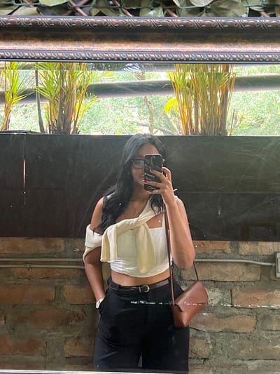A young woman takes a mirror selfie. She wears dark pants, a light top knotted at the waist, and glasses. A brown purse hangs from her shoulder. The mirror is framed with a decorative gold-colored border. The reflection shows greenery through a window on one side and a brick wall on the other. The flash reflects on the mirror.