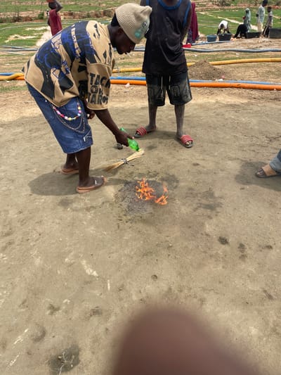 A ground-level shot, rotated to a horizontal orientation, captures a scene in Maiyanga, Nigeria. The ground is pale and dry. To the left, irrigation pipes are visible, indicating an agricultural setting. Two people are present. One man wears a beige beanie, a beige t-shirt with blue and black designs, blue jeans, and tan sandals; he crouches and holds a green bottle and a bundle of twigs over a small fire burning on the ground. A second individual, partly visible, wears a black shirt, black shorts, and red striped sandals. The fire is the central focus, with flames rising from a dark, ashy base.
