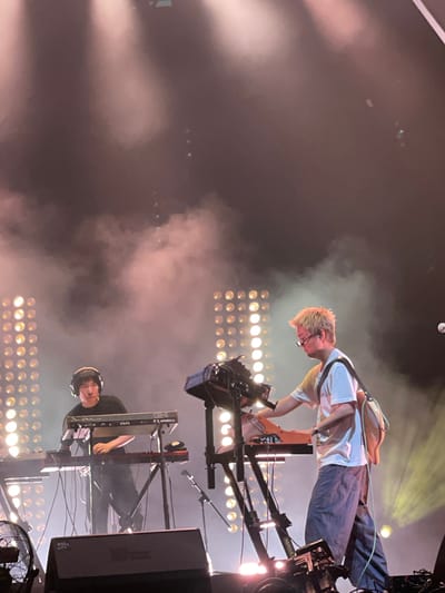 The image shows two individuals performing on a stage. The individual on the right side is wearing a white t-shirt, blue pants, and eyeglasses, and is bending over, possibly playing a musical instrument. The individual on the lower side is seated and wearing headphones, with their hands on a keyboard. The stage is equipped with multiple light fixtures and sound equipment. Based on the image content, the location is not possible to determine.