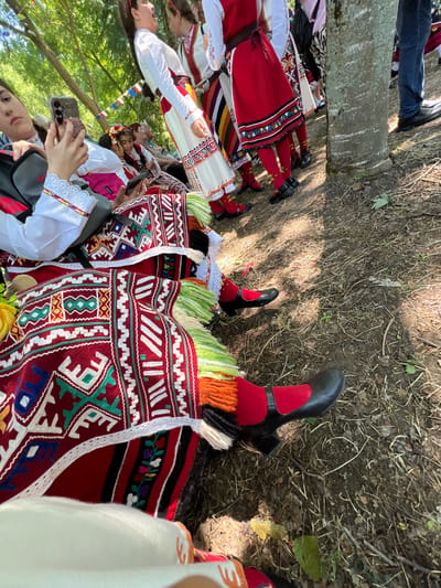 This image depicts an outdoor scene in Bolyarovo, Bulgaria, featuring multiple individuals in traditional Bulgarian folk costumes. In the immediate foreground on the left, a young woman is seated, holding and looking at a smartphone with a gold/rose gold frame and a three-camera array. She wears a white long-sleeved blouse with lace cuffs and red/yellow trim, a dark vest, a red apron-style garment with intricate geometric and floral patterns in white, green, blue, yellow, red, and black, and has vibrant green and orange fringed tassels extending from her costume. Her leg is visible, showing a red stocking and a black Mary Jane-style shoe with a strap and heel. A gray Nike backpack rests next to her.
To her right, partially obscured, another person's foot is visible, also wearing a red stocking and a black shoe. Further back, several other individuals are seated and standing. Another young woman with a floral headpiece (red and yellow flowers) is seated, also looking at a smartphone. An older woman in a white blouse and dark attire is seated behind her.
In the middle ground, several women stand wearing variations of traditional costumes, characterized by white blouses, colorful striped or red skirts/aprons with embroidered bands at the hem, often featuring floral or geometric motifs. Red stockings and black Mary Jane-style shoes are common footwear. One woman wears a white blouse, a red apron with multi-colored embroidery, and a vertically striped skirt. Another has a white blouse and a vibrant striped skirt.
On the far right, a large tree trunk with rough bark is prominent. Behind it, the lower legs of a man wearing blue jeans and black dress shoes are visible.
The ground consists of dry earth, scattered dry leaves, twigs, and sparse patches of green vegetation, indicating an outdoor, possibly shaded, environment. Overhead, the canopy of trees is visible. In the background, strung between trees, a line of colorful rectangular bunting flags (red, white, blue, green, yellow) suggests a festive event. The scene is illuminated by dappled sunlight, creating areas of light and shadow on the ground.