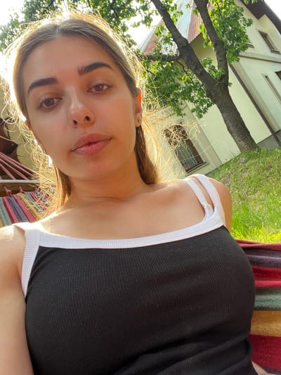 The image shows a young woman lying down outdoors on a striped hammock, with her head tilted towards the camera. She is wearing a black tank top with white trim. Sunlight filters through the trees and grass behind her, suggesting it is daytime. The background shows part of a building and some greenery, indicating an outdoor setting, possibly a backyard or garden. The woman's expression is relaxed, with her eyes open and looking at the camera. Her hair is long and appears to be brown. The overall impression is a casual, sunny day.
