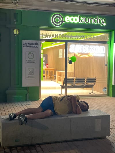 The image shows a man sleeping outdoors next to a laundromat called "ecolaundry". The laundromat has a prominent green and white sign. Inside the laundromat, neon signs for "Amazon" and "Amazon Hub" are visible, suggesting it might also serve as a drop-off or pick-up point for Amazon packages. The man is wearing a yellow t-shirt and blue shorts, with his arms crossed as he leans against a large concrete pillar. His shoes are visible, and he appears to be sleeping soundly. The scene is set at night, indicated by the artificial lighting from the laundromat and the darkness of the surroundings. The brick wall of an adjacent building is also visible. The laundromat also displays information in Spanish, including "LAVANDERÍA AUTOSERVICIO", "EN TAN SOLO 1 HORA LAVA Y SECA TODO TIPO DE ROPA", and "MÁQUINAS DE GRAN CAPACIDAD". The presence of the Amazon Hub signage suggests the location is likely in an urban or semi-urban area. The overall impression is one of an unusual overnight resting place for the man, juxtaposed with the functionality of the laundromat and Amazon services.