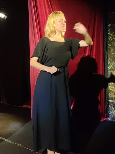 A blonde woman in a black dress stands on a stage in Bedulu, Indonesia, near a red curtain. She is gesticulating with her right hand, and a shadow falls behind her on a patterned wall. A stage light illuminates her from the left.