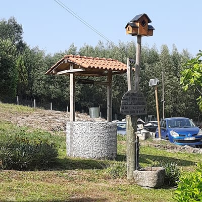 The image depicts an outdoor scene in Vila Verde, Portugal, featuring a traditional well as the central element. The well has a stone-lined circular base and a wooden structure above with a tiled roof, supporting a rope and bucket for drawing water. To the right of the well, a wooden post displays a carved sign with Portuguese text that reads: "A ÁGUA É FONTE DE VIDA / A VIDA É FONTE DE AMOR / SE A FONTE FICA POLUÍDA / A VIDA SOFRE DE AMOR". Adjacent to the post is a small solar panel mounted on a wooden pole, likely powering a light. Further to the right, a blue car is parked on a grassy area, and behind it, a dense line of trees forms the background. The ground is covered with grass and dry earth, with some vegetation in the foreground. The sky is clear and blue.