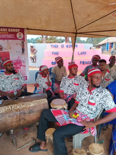 An outdoor cultural gathering is depicted under a light-colored tent, providing shade from the bright daytime sun in Omicha Umu Awada, Nigeria. Approximately 10-12 men, mostly young adults, are seated on plastic chairs. Most are dressed in traditional attire: white and black patterned short-sleeved shirts with red trim, and red and white striped traditional caps. Many also wear plaid wrappers tied around their laps.
Prominently featured are traditional musical instruments, including a large wooden drum, likely a slit drum or ekwe, with "MMANWU OPALA" carved into its side. Several smaller traditional drums are also visible on the ground. A smartphone rests on the large drum on the left, and some tools are on the ground nearby.
Behind the seated group, a pink tiered structure serves as a backdrop, with blue text reading "THIS IS EZIO", "THE LAND", and "OF PEACE". To the left, a banner displays text such as "LADE GROUP", "COMMUNITY", and "STATE, NIGERIA," alongside the Nigerian coat of arms and images of traditionally dressed people. The ground under the tent is dirt or sand. The men appear calm and attentive, some looking towards an unseen focal point.