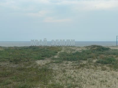 The image shows a sandy landscape with sparse vegetation leading to the sea. In the background, large white letters spelling "КРАЙ ЗЕМЛИ" (Kray Zemli, meaning "Edge of the Earth") stand on a slight rise, with the sea and sky visible behind them. A blue pole with a horizontal bar is visible on the far right edge of the frame. The scene depicts a coastal area in Yamburg, Russia, likely a landmark indicating the extremity of the land. There are no people visible in the image. The environment is open and natural, characterized by sand dunes and sea grass.