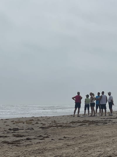 The image captures a group of six individuals standing on a sandy beach in South Padre Island, United States of America, under an overcast, light gray sky. The foreground consists of light brown sand with scattered clumps of dark, organic debris. To the left, the ocean displays moderate waves with visible whitecaps extending towards a hazy horizon. The individuals, all appearing to be adults, are positioned in a loose cluster in the mid-ground, oriented towards the ocean. From left to right, the group includes: a male in a red sleeveless top and dark shorts with hands on hips; an individual in a light green shirt and dark shorts; a female in a light-colored top and dark shorts; a male in a light blue t-shirt and dark shorts; a male in a white sleeveless top and dark shorts; and a male in a light-colored collared shirt, dark shorts, and a light-colored baseball cap, with his hands clasped behind his back. No specific interactions between the individuals are evident.