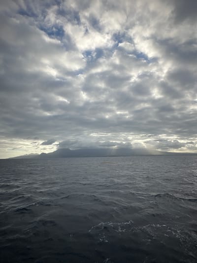 The image depicts a wide expanse of choppy, dark blue ocean under a cloudy sky. Shafts of sunlight pierce through gaps in the clouds, illuminating parts of the ocean and casting shadows. In the distance, a silhouetted mountain range forms a landmass across the horizon. One small, elongated yellow object is visible floating on the water's surface, to the left of the mountain range. The scene is likely a view from a boat off the coast of Capesterre-Belle-Eau, Guadeloupe, given the presence of volcanic mountains characteristic of the region and the surrounding ocean.