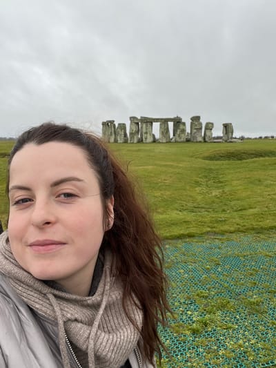 The image displays a selfie-style photograph featuring an adult female in the foreground against the backdrop of Stonehenge, located near Larkhill, Wiltshire, United Kingdom. The time is daytime under an overcast sky, casting a soft, diffused light.
The female subject, positioned prominently in the lower-left portion of the frame, has fair skin, dark brown hair pulled into a ponytail, and a slight, closed-mouth smile. She wears a light-colored (beige or light grey) hooded, ribbed knit neck warmer or cowl over a darker garment, topped by a light grey puffer jacket. A small silver stud earring is visible in her left earlobe. Her gaze is directed slightly to the viewer's right.
In the mid-ground, the ancient monument of Stonehenge is visible. It comprises numerous large, grey, weathered sarsen stones, some standing upright and others forming trilithons with horizontal lintel stones. The monument is situated on an expansive, undulating green grassy plain. The immediate foreground shows a section of green plastic or mesh ground reinforcement partially covered by grass. In the distant background, beyond Stonehenge, the horizon is flat, and faint, indistinct human figures can be observed near the monument. The overall environment is open and unpopulated except for the monument and a few distant individuals.