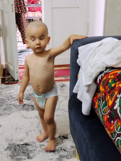 A bald toddler, wearing a diaper, stands barefoot on a patterned carpet indoors. The child is positioned near a dark blue sofa adorned with a colorful, patterned fabric. To the left of the child, a white door is slightly ajar, revealing a glimpse of a doorway with patterned curtains and shelving. The overall scene suggests a domestic setting within a home in Namangan City, Uzbekistan.
