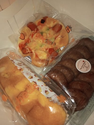 Three distinct packaged baked goods are displayed on a light-colored surface in Khajuripada, India. A round savory bun, topped with sliced sausages, melted cheese, and diced vegetables, is sealed in clear plastic marked "HALAL". Adjacent to it, a rectangular golden-brown sweet bread loaf, garnished with almond flakes, is also encased in clear plastic featuring a decorative paper strip with cupcake motifs and the "HALAL" designation. To the right, a clear plastic container holds approximately ten dark brown, individual ring-shaped cakes, identified by a circular sticker as "Kue Cincin Kopoor Gula Merah". No individuals are visible in the static scene.